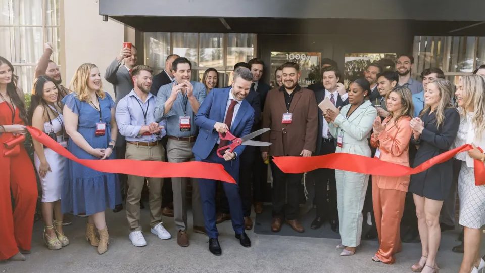 Mac Clemmens cutting a red ribbon with oversized scissors at the opening of the Streamline office, surrounded by a cheering team.
